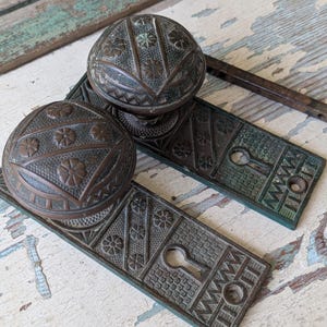 May include: Antique bronze door hardware set featuring two ornate doorknobs and matching backplates. The doorknobs have a rounded shape with floral and geometric patterns. The backplates have keyholes and decorative detailing, showcasing a vintage aesthetic.