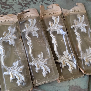 May include: Four vintage white and gold cabinet pulls in their original packaging. The pulls have an ornate, floral design. The packaging reads "Quality Cabinet Accessories" and "PULL". The pulls are made by National Lock.