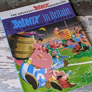 May include: A blue and white comic book cover featuring the title "The Greatest Asterix Adventure Asterix in Britain by Goscinny and Uderzo". The cover features a cartoon illustration of Asterix and Obelix playing rugby.