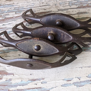 May include: Four antique, dark brown metal drawer pulls. Each pull has a central oval plate with a rounded knob and decorative openwork ends. The pulls are arranged in a slightly overlapping pattern, showcasing their aged appearance.