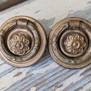 May include: Two antique brass drawer pulls. Each pull features a circular design with a central floral motif and a hinged ring handle. The pulls have an aged, tarnished appearance, suggesting vintage or reclaimed hardware.