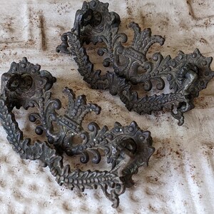 May include: Two ornate metal drawer pulls with a decorative, floral design. The pulls are a dark, aged metal color.