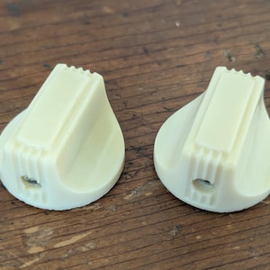 May include: Two vintage cream-colored guitar knobs. Each knob has a rectangular top with a ridged edge and a teardrop-shaped base. The knobs are made of a solid, opaque material and are designed for use on a guitar or other musical instrument.