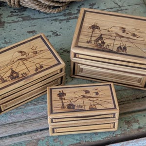 May include: Three decorative wooden boxes with etched landscape designs. The boxes are rectangular with a light brown color and darker brown trim. The lids feature etched images of palm trees, buildings, and clouds. The boxes are arranged on a weathered, teal-colored wooden surface.