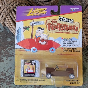 May include: A Johnny Lightning die-cast metal toy car of Barney Rubble's sports car from The Flintstones cartoon series. The packaging features images of Fred and Barney in their car, and the text "Main St."