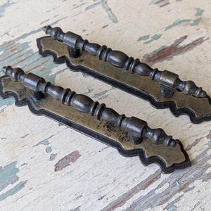 May include: Two matching drawer pulls featuring a dark bronze finish. Each handle has a central rod with decorative turned detailing, mounted onto a flat, scalloped-edge backplate with a weathered, aged brass appearance and dark edges.
