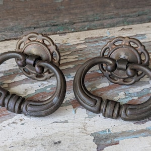 May include: Two antique bronze drawer pulls with ornate details. Each pull features a circular design with a raised edge and a loop handle.