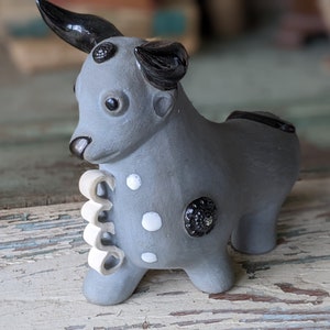 May include: A gray ceramic bull figurine with black horns and eyes. The bull has white and black spots and a white curly tail.