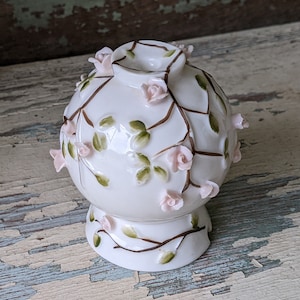 May include: A spherical white porcelain vase featuring a raised floral design with small pink sculpted roses and hand-painted green leaves on brown vines. The vase has a circular opening at the top and a flared base, with the floral pattern extending down to the bottom.