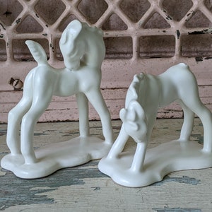 May include: Two white ceramic figurines of horses, one standing and the other lying down. Both horses have a glossy finish and are posed in a playful manner.