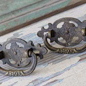 May include: Two antique bronze drawer pulls with ornate floral designs. Each pull features a decorative backplate and a curved handle. The pulls are set against a weathered, light-colored wooden surface, showcasing their vintage aesthetic.