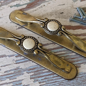 May include: Two antique brass cabinet pulls with ornate designs and a white center. The pulls have a decorative, floral-like design.