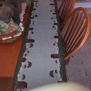 May include: A long, rectangular table runner featuring a light blue center panel bordered by a dark green edge. The border is decorated with a repeating pattern of small red barns and green trees. The fabric has a woven texture throughout the design.