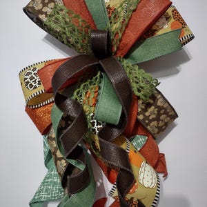 May include: A decorative bow featuring a variety of ribbons in fall colors. The bow includes ribbons with pumpkin and leopard print patterns, solid orange, green, and brown ribbons, and a brown bow center. The bow is suitable for fall and autumn decor.
