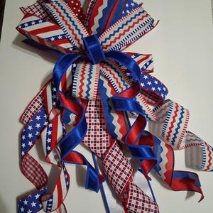 May include: A decorative bow featuring red, white, and blue ribbons with various patterns, including stars, stripes, polka dots, and zigzags. The bow is tied with a blue satin ribbon and is suitable for patriotic or holiday decorations.