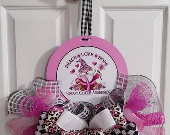 Puede incluir: Corona rosa y negra con un lazo de cinta rosa y una cinta rosa y blanca con estampado de leopardo. La corona tiene un círculo rosa en el centro con un gnomo que lleva un sombrero rosa y las palabras "Peace Love Hope" y "Breast Cancer Awareness".