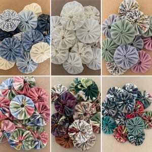 May include: A selection of fabric yo-yos in diverse colours and patterns. These round yo-yos have a gathered centre and are made from fabrics such as lace, floral prints, and solid colours. The colour scheme includes blues, pinks, greens, and creams.