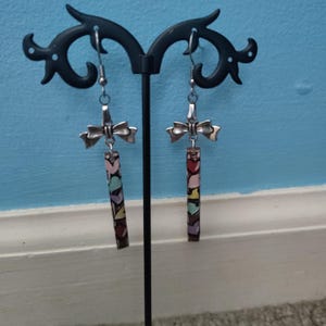 May include: A pair of dangle earrings with a bow-shaped top and a rectangular pendant. The pendant is decorated with colorful heart shapes. The earrings are displayed on a black earring stand against a blue wall.