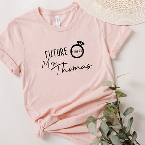 May include: A pink t-shirt with the text "FUTURE Mrs Thomas" and a ring with a diamond and the date "21.06.22" printed on it.
