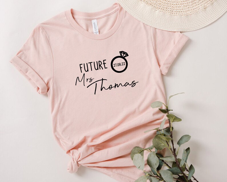 May include: A pink t-shirt with the text "FUTURE Mrs Thomas" and a ring with a diamond and the date "21.06.22" printed on it.