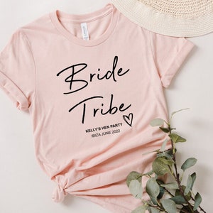 May include: Pink t-shirt with the text "Bride Tribe" in black script font and a small heart. Below the text is "Kelly's Hen Party Ibiza June 2022" in black.