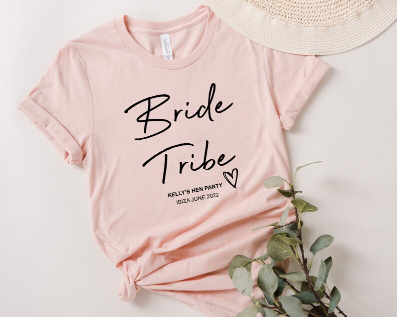 May include: Pink t-shirt with the text "Bride Tribe" in black script font and a small heart. Below the text is "Kelly's Hen Party Ibiza June 2022" in black.