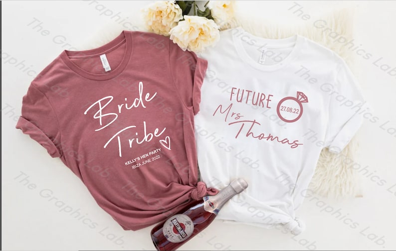 May include: Two white t-shirts with text printed on them. The shirt on the left says "Bride Tribe" with a heart and the date "08.24.JUNE 2022". The shirt on the right says "FUTURE Mrs Thomas" with a ring and the date "27.08.22".