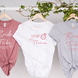 May include: Three t-shirts with text printed on them. The first t-shirt is pink and says "Bride Tribe" with the date "18th June 2022". The second t-shirt is white and says "Future Mrs. Thomas" with the date "27.08.22". The third t-shirt is grey and says "Bride Tribe" with the date "18th June 2022".
