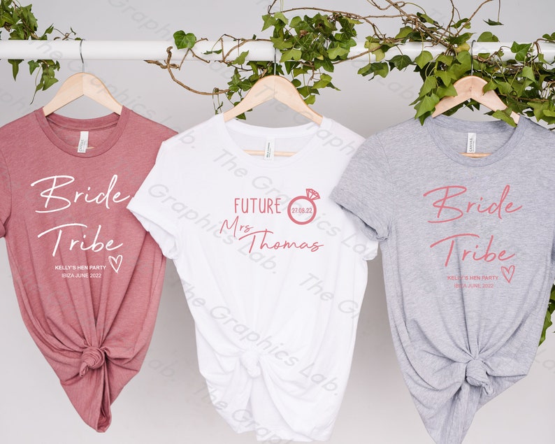 May include: Three t-shirts with text printed on them. The first t-shirt is pink and says "Bride Tribe" with the date "18th June 2022". The second t-shirt is white and says "Future Mrs. Thomas" with the date "27.08.22". The third t-shirt is grey and says "Bride Tribe" with the date "18th June 2022".