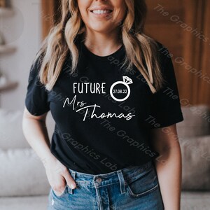May include: Black t-shirt with white text that reads "FUTURE Mrs Thomas" and a white ring with a diamond graphic and the date "27.08.22".