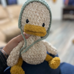 May include: A handmade crochet duck plushie with a light beige body, a yellow beak and feet, and black eyes. The duck is wearing a light green bonnet. The plushie is soft and cuddly, perfect for a child's toy or nursery decor.