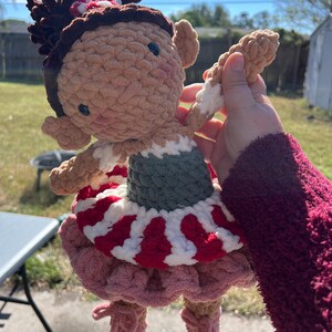 May include: A handmade crochet doll with a light brown face, dark brown hair, and blue eyes. The doll wears a green and white top, a red and white striped skirt, and a pink ruffled skirt. The doll is holding a tan and white object.