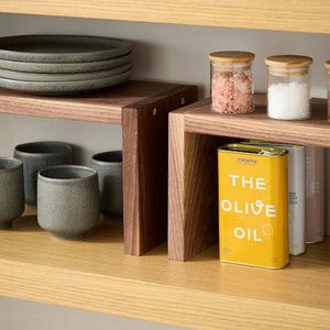 Op de afbeelding: Een houten keukenkast met grijze keramische borden, bekers en glazen potten met houten deksels. Een gele blik olijfolie met de tekst "THE OLIVE OIL" is ook zichtbaar. De plank is gemaakt van licht en donker hout.