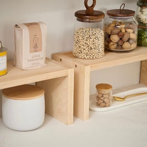 Op de afbeelding: Een keuken met lichte houten planken met diverse items. Een koffiezak, glazen potten met houten deksels gevuld met bonen en noten, en een witte container met een houten deksel zijn zichtbaar. Een klein potje met een houten deksel en een gouden lepel zijn ook aanwezig.