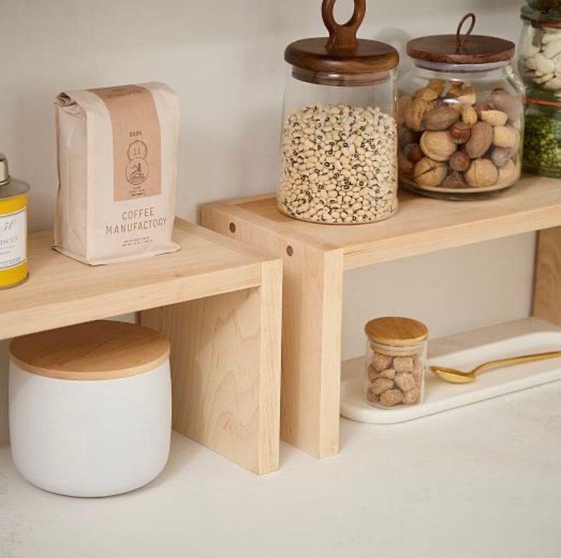 Op de afbeelding: Een keuken met lichte houten planken met diverse items. Een koffiezak, glazen potten met houten deksels gevuld met bonen en noten, en een witte container met een houten deksel zijn zichtbaar. Een klein potje met een houten deksel en een gouden lepel zijn ook aanwezig.