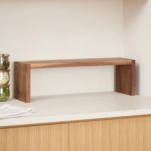 Op de afbeelding: Een houten plank met een rechthoekige bovenkant en twee verticale steunen. De plank is gemaakt van donkerbruin hout en staat op een wit aanrechtblad. De plank is ongeveer 61 cm lang.