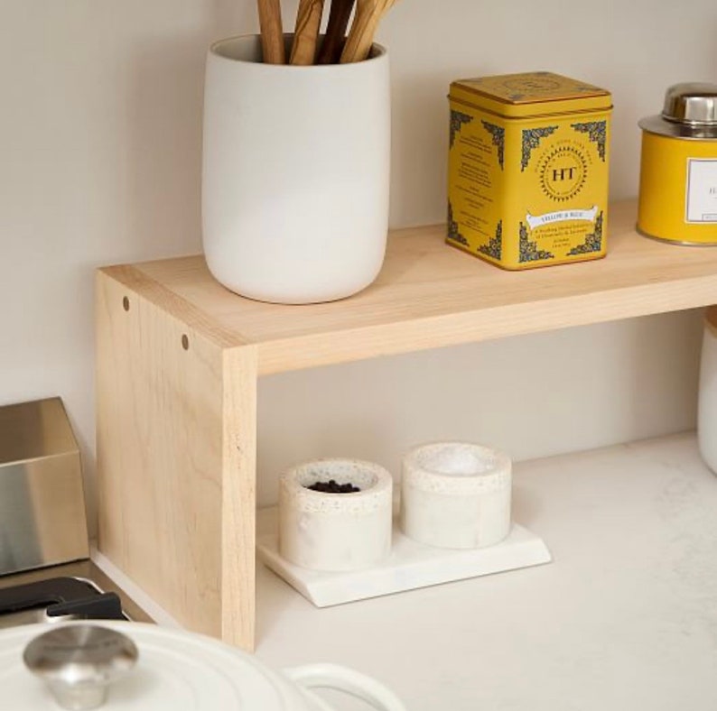 Op de afbeelding: Een lichtgekleurd houten plank met een witte keramische keukengerei houder, een gele theeblik en kleine witte bakjes. De plank wordt ondersteund door een zijpaneel. De plank staat op een wit aanrecht.
