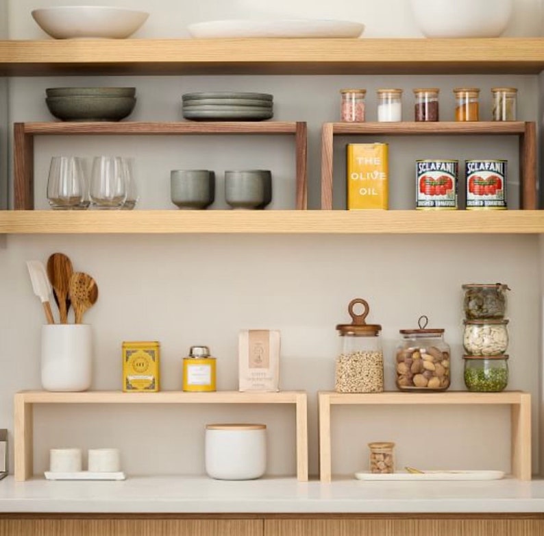 Op de afbeelding: Een keukenrek met diverse items. Houten planken bevatten kommen, glazen, kruidenpotjes, conserven en andere keukenspullen. Het kleurenpalet is neutraal, met houttinten en witte accenten.