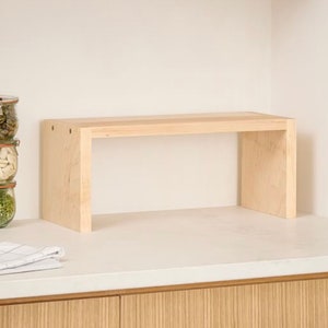 Op de afbeelding: Een lichtgekleurd houten plank, rechthoekig van vorm, met een platte bovenkant en twee zijsteunen. De plank staat op een wit aanrechtblad, met potten en een gevouwen doek zichtbaar op de achtergrond. De plank is ongeveer 30 cm hoog en 61 cm breed.