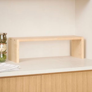 Op de afbeelding: Een lichtgekleurd houten plank met een rechthoekig ontwerp. De plank wordt ondersteund door twee verticale panelen, waardoor een eenvoudige, minimalistische esthetiek ontstaat. De plank is ontworpen om op een aanrecht te staan. Het hout heeft een natuurlijke afwerking.