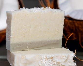 May include: Three bars of handmade soap stacked on top of each other. The top bar is white with a light gray layer at the bottom. The middle bar is white with a light gray layer at the bottom. The bottom bar is light gray. The soap is decorated with white coconut flakes.