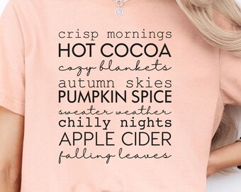 May include: A peach-colored t-shirt with the words "crisp mornings, HOT COCOA, cozy blankets, autumn skies, PUMPKIN SPICE, sweater weather, chilly nights, APPLE CIDER, falling leaves" printed in black. The shirt is paired with blue jeans.