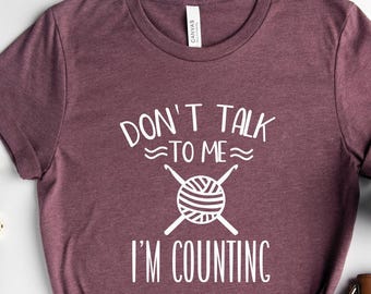 May include: Heather maroon t-shirt with the text "DON'T TALK TO ME I'M COUNTING" in white. The graphic includes a ball of yarn and knitting needles. Accessories include a watch, glasses, earrings, a coffee mug, and a brown purse.