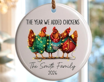 May include: A round white ceramic ornament hanging from a brown twine string. The ornament features an illustration of three chickens wearing colorful sweaters and string lights, with the text "THE YEAR WE ADDED CHICKENS", "The Smith Family", and "2026".