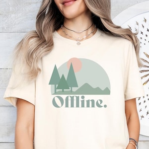 May include: Cream-colored t-shirt featuring a nature-themed graphic with trees, a mountain, and a sun. The word "Offline" is printed below the graphic in a sage green color. The shirt is made of a soft material.