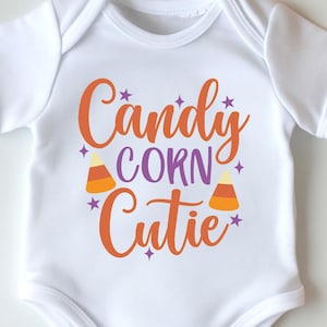 May include: White baby onesie with the text "Candy Corn Cutie" in orange and purple. The onesie is surrounded by pumpkins and fall leaves, creating a festive Halloween theme.