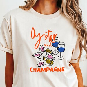 May include: Beige t-shirt with the words "Oyster and Champagne" in orange script. The shirt features a graphic of oysters, lemon slices, and two wine glasses with blue liquid.