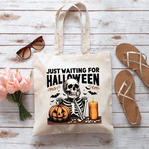 May include: Beige tote bag with a Halloween-themed graphic. The design features a skeleton, a jack-o'-lantern, a lit candle, and the text "JUST WAITING FOR HALLOWEEN". The bag is on a wooden surface, with sunglasses, flowers, and sandals.
