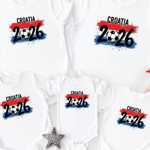 May include: White t-shirts with the text "CROATIA 2026" above a soccer ball graphic. The design features the Croatian flag colors: red, white, and blue. The shirts are available in various sizes, including a baby onesie.
