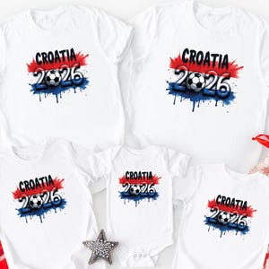 Puede incluir: Camisetas blancas y un body con un dise&ntilde;o de f&uacute;tbol Croacia 2026. El dise&ntilde;o presenta la palabra "Croacia" en negro, un bal&oacute;n de f&uacute;tbol y el a&ntilde;o "2026" en azul, con salpicaduras de pintura roja y azul.